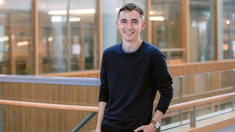 UQ Student Jasper Stead stands smiling in front of a banister with glass windows in the background