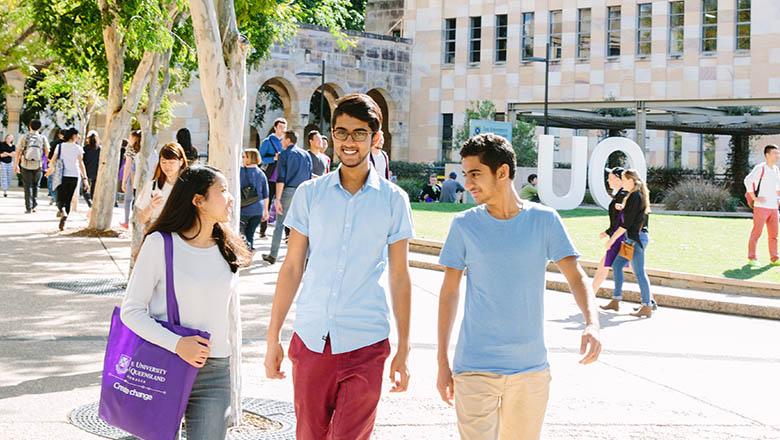UQ students walk and talk to one another with sandstone buildings and trees in the background