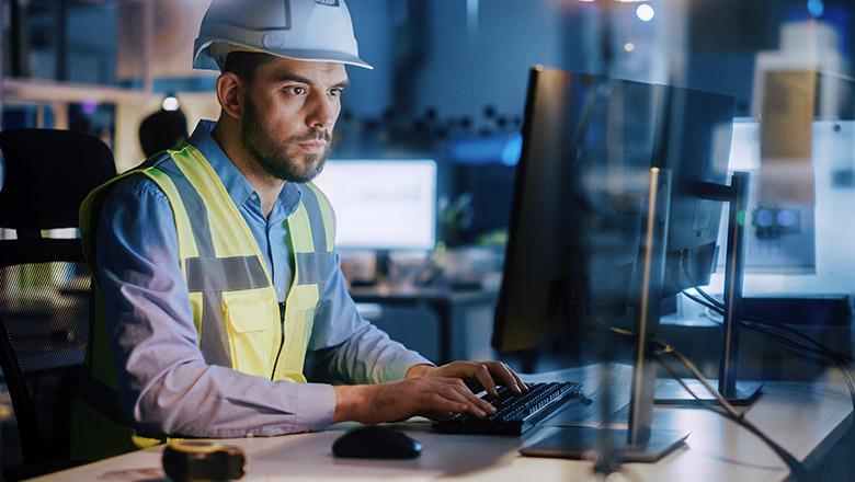 An employee in a hard hat and hi-vis vest sits at a desk typing on a computer