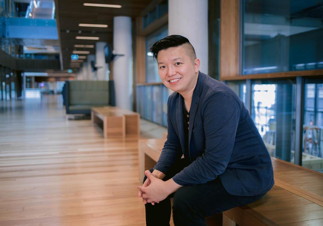 Professor Ryan Ko sits with hands interlinked and smiling on a wooden bench indoors