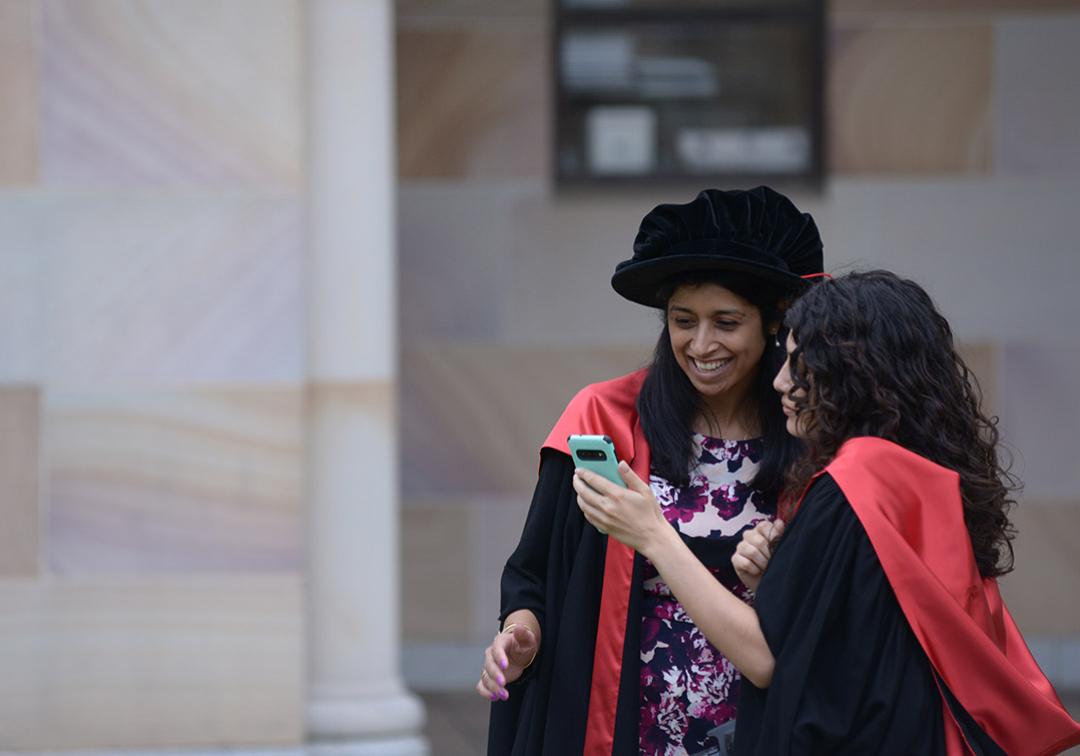 Doctoral graduate in gown and graduation tam, looking at phone with friend