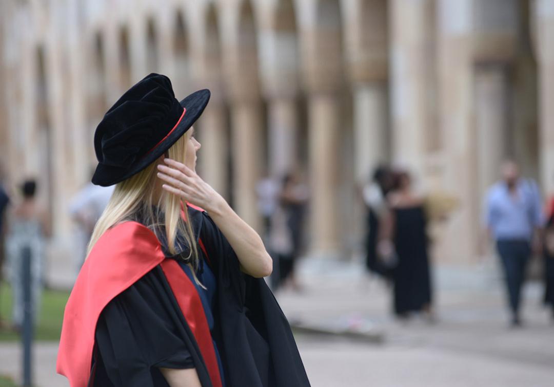 Blonde woman in a graduation tam and robe sweeps her hair from her face, with sandstone university blurred in background