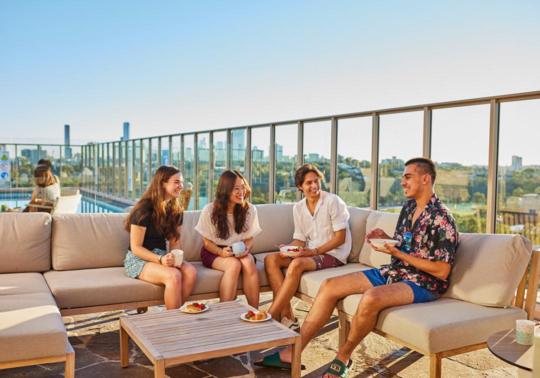 Students sit on rooftop pool deck talking and laughing, with cityscape in the background