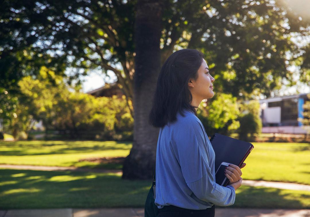 Female student walks holding books and phone, with sunlit tree and grass in background