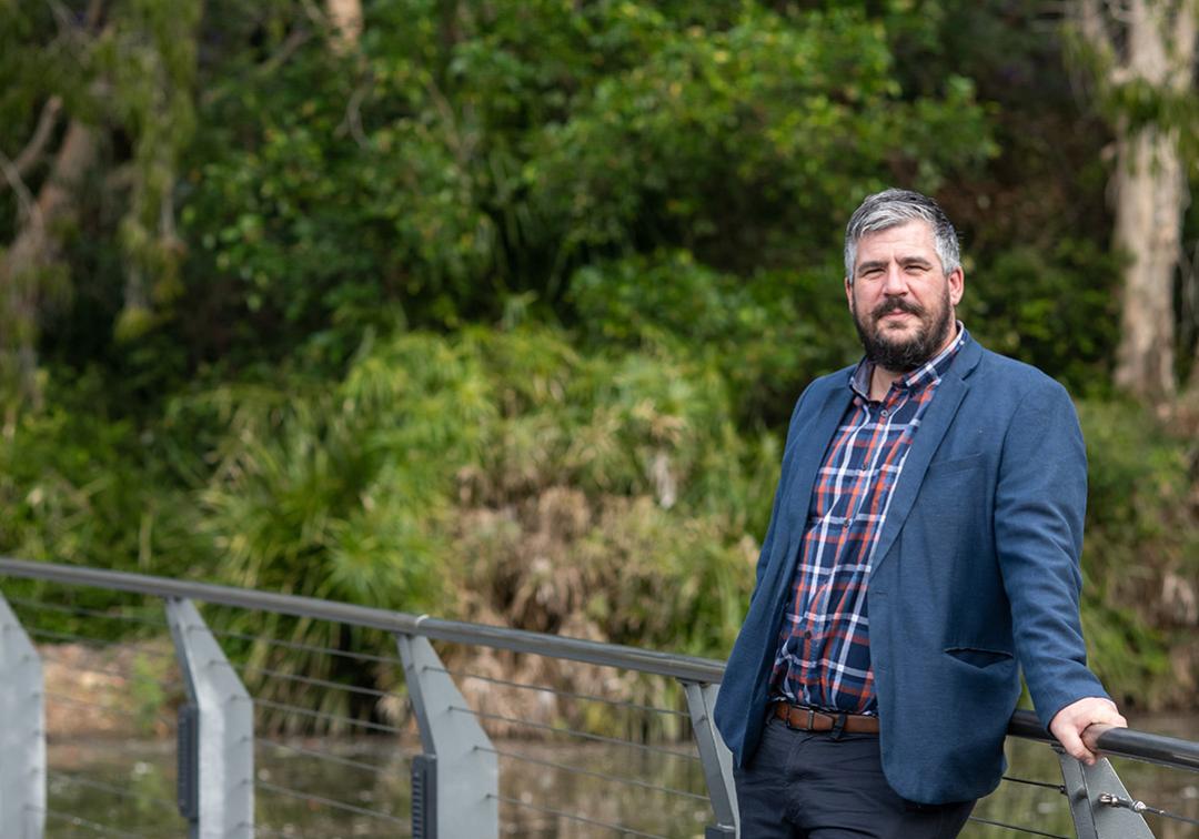 Dr Alastair Stark leans against the railing at UQ Lakes with green vegetation in background