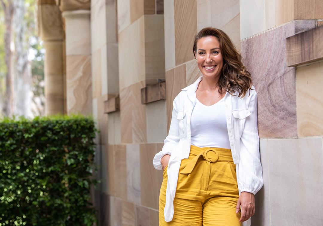 Natalie Craig stands smiling, leaning against UQ's sandstone buildings