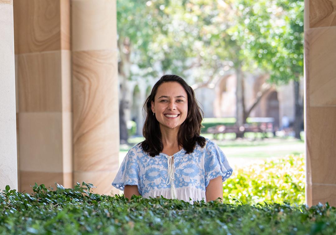 Barbara Azevedo de Oliveira stands smiling in UQ's Great Court, flanked by sandstone and trees