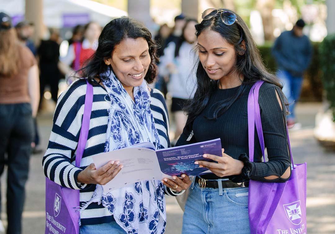 A mother and daughter at UQ Open Day looking at a booklet and holding purple UQ tote bags