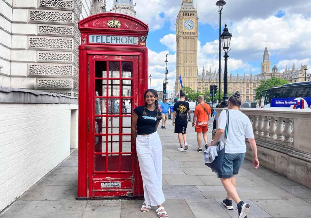 Esandi Kalugalage stands in front of a red phone box on a street in London with Big Ben in the background