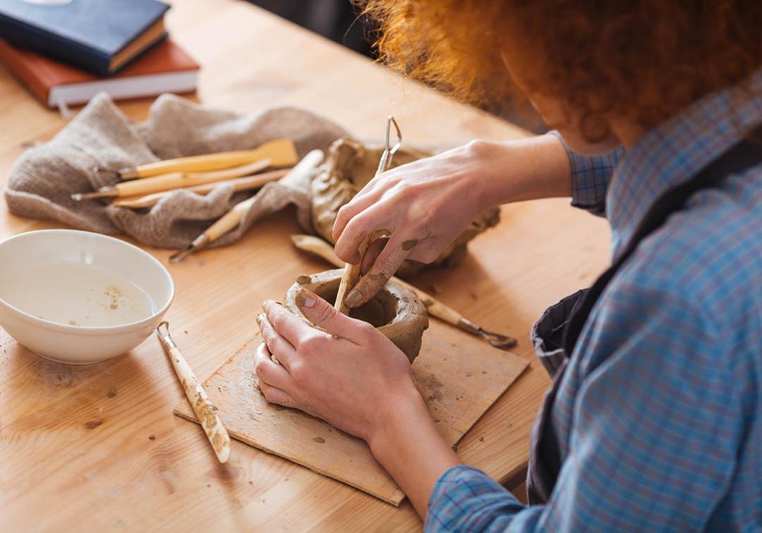 A person leans over clay, which they are shaping into a bowl with tools