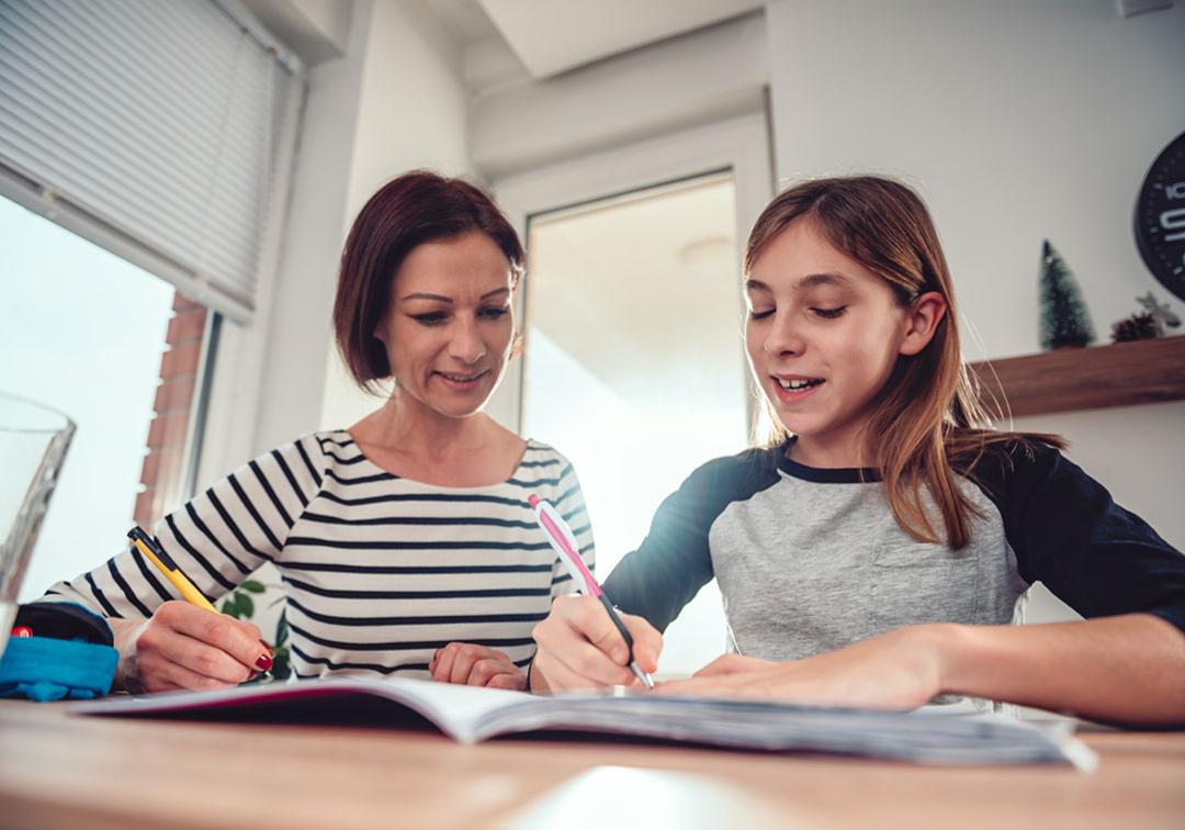 Mother and daughter sit at desk holding pens and looking over notebook