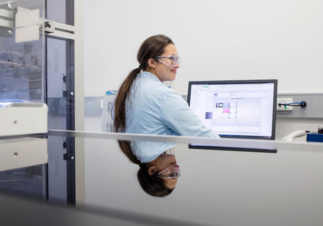 A UQ student in a lab coat and safety glasses sits in front of a computer in a lab