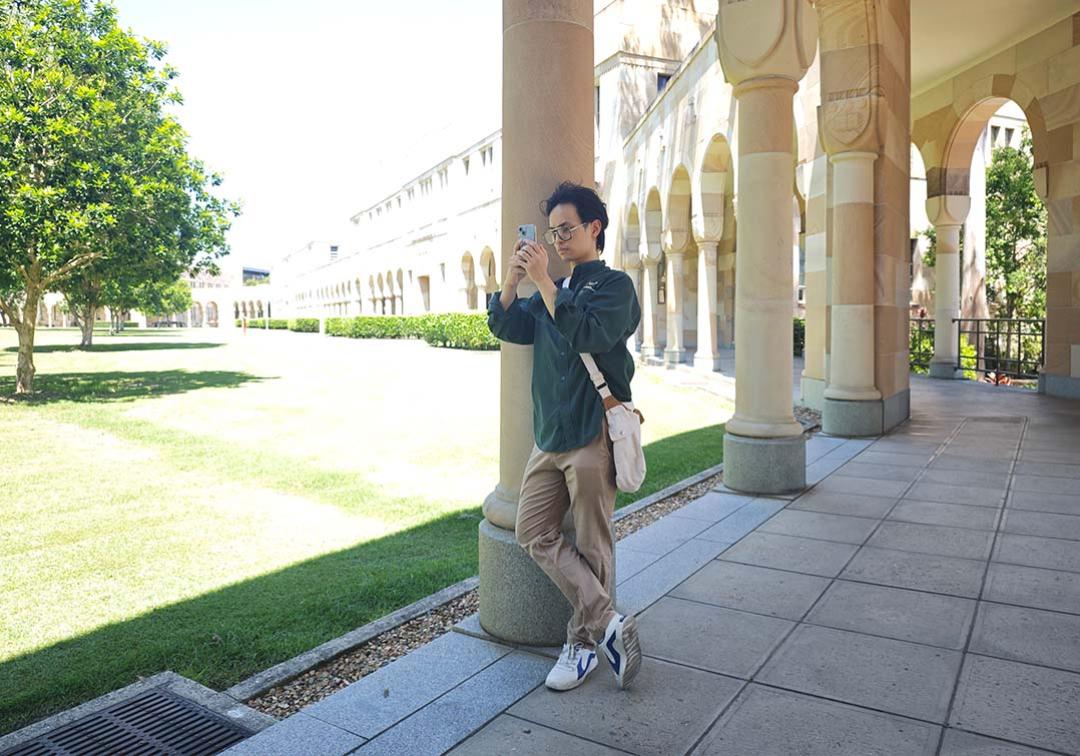 Male student leaning on a cloister taking a photo with his phone