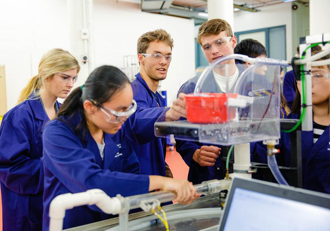 A group of UQ engineering students in lab coats and safety glasses inspect an experiment