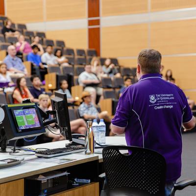 Man in purple UQ t-shirt speaking to a lecture theatre of students
