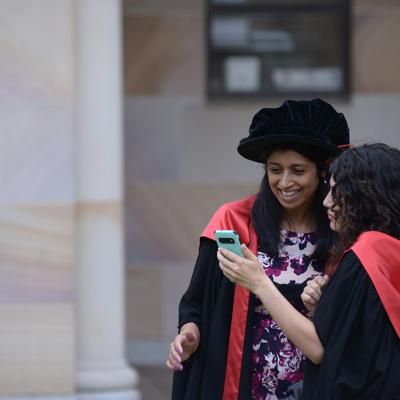 Doctoral graduate in gown and graduation tam, looking at phone with friend