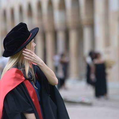 Blonde woman in a graduation tam and robe sweeps her hair from her face, with sandstone university blurred in background