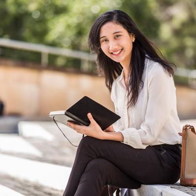 A young woman holding a tablet sits outside, smiling while looking at the camera