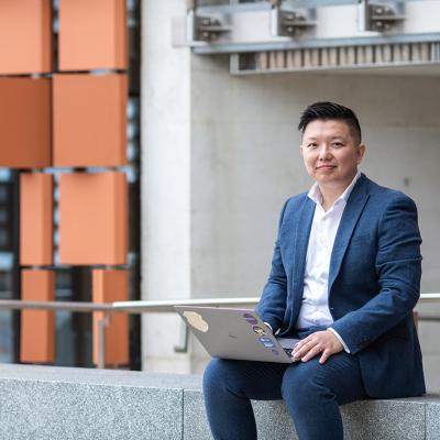Professor Ryan Ko sitting with laptop in front of concrete building
