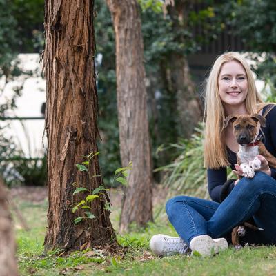 Zoë Black sits crossed legged holding a puppy  and smiling with trees and greenery in the background