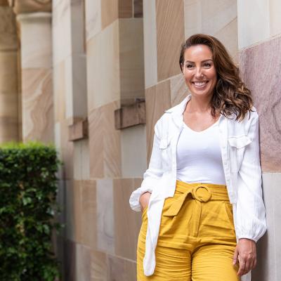 Natalie Craig stands smiling, leaning against UQ's sandstone buildings