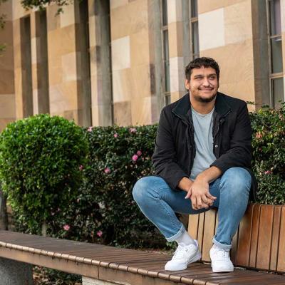 UQ student Jamaine Wilesmith sits in The Great Court with shrubbery and sandstone buildings in the background