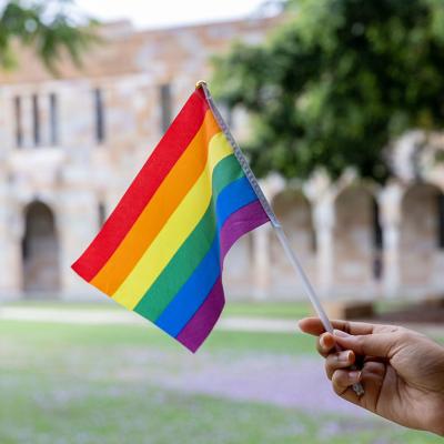 a hand holds a rainbow flag in front of UQ St Lucia's sandstone cloisters