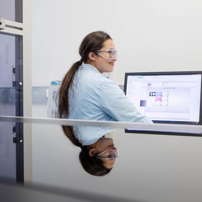 A UQ student in a lab coat and safety glasses sits in front of a computer in a lab