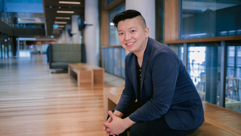 Professor Ryan Ko sits with hands interlinked and smiling on a wooden bench indoors