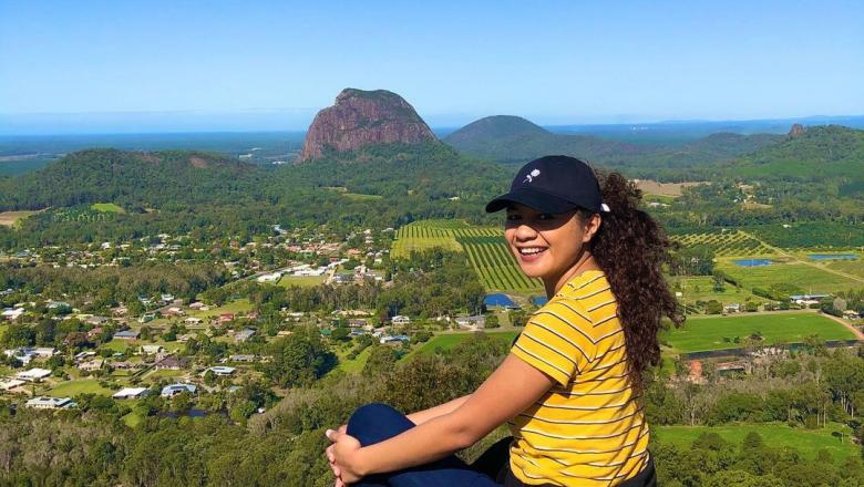 International student sat on rock with views of glasshouse mountains, green fields and blue sky behind her