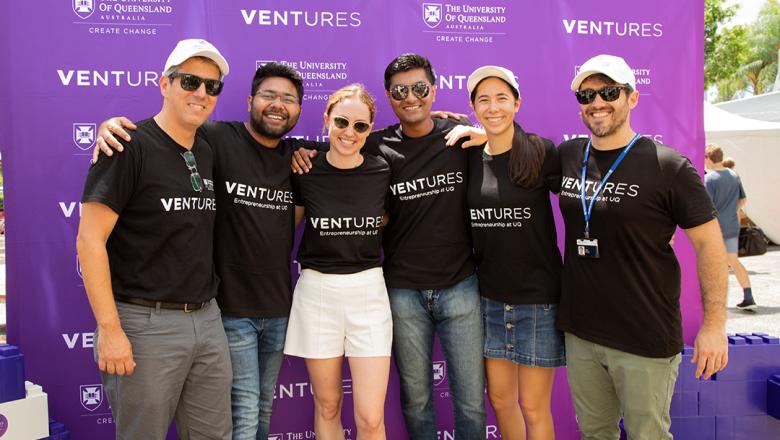 Six students in black UQ Ventures tshirts have their arms around each other while standing infront of a purple UQ Ventures banner