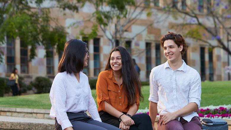 Three international students sat on wall infront of green grass and stone walls at UQ St Lucia
