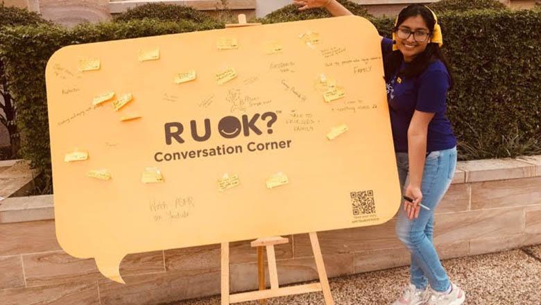 UQ Student Megha Thakkar is wearing jeans and a t-shirt, displaying a large yellow posterboard in the shape of a speech bubble with the words 'R U OK? Conversation Corner' and post-it notes stuck onto it
