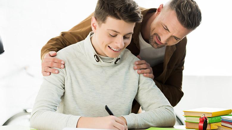 A Father leans over his seated son smiling as he sees what he has written in his school homework