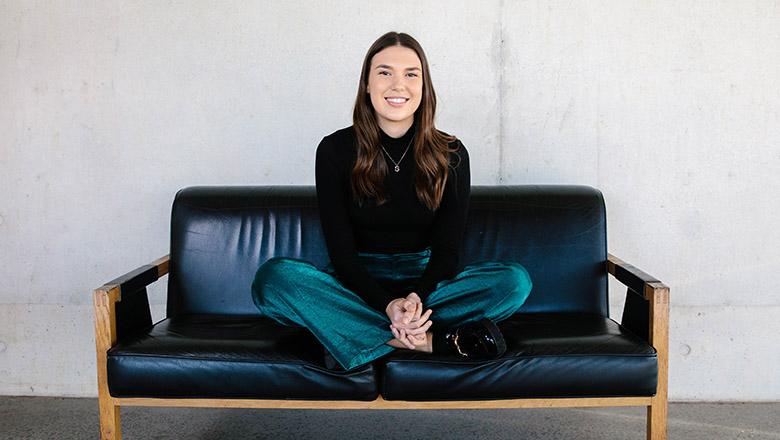 Indigenous studies students Sylvia Stuen-Parker sits cross-legged on a black leather couch, smiling at the camera