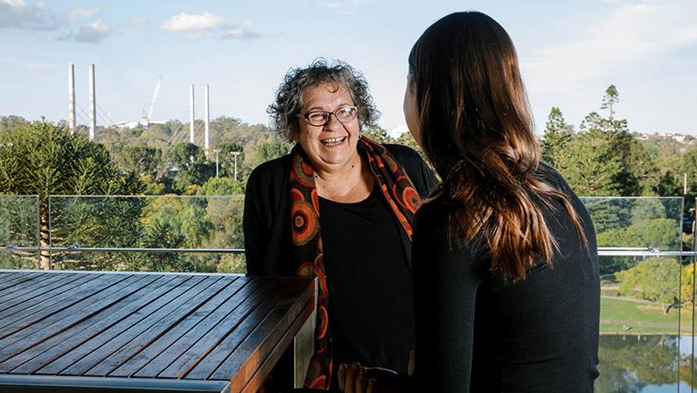 Tracey Bunda sits smiling at Sylvia Stuen-Parker as she talks, with the UQ St Lucia campus and Eleanor Schonell Bridge in the background