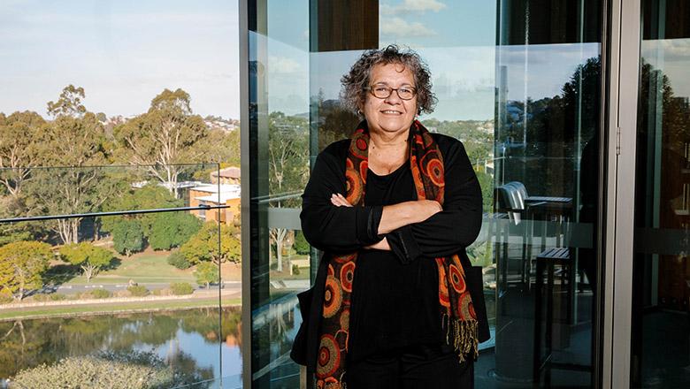 Indigenous studies Professor Tracey Bunda stands smiling at the camera with her arms crossed, in front of glass walls that look out over UQ St Lucia campus