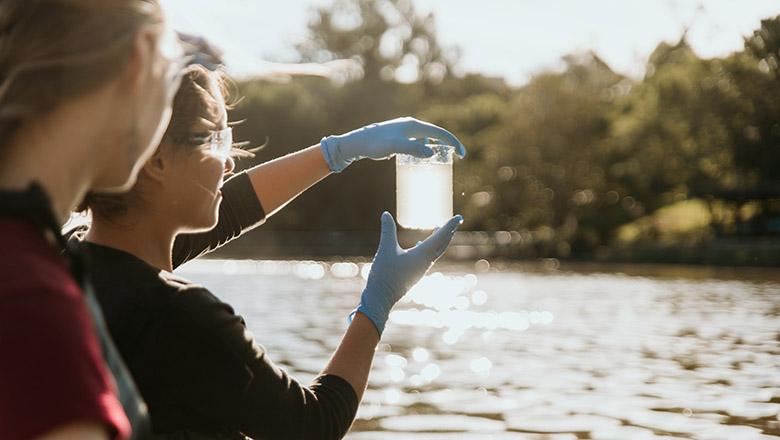 A water quality specialist holds a test tube of water up to the light to inspect with a lake in the background