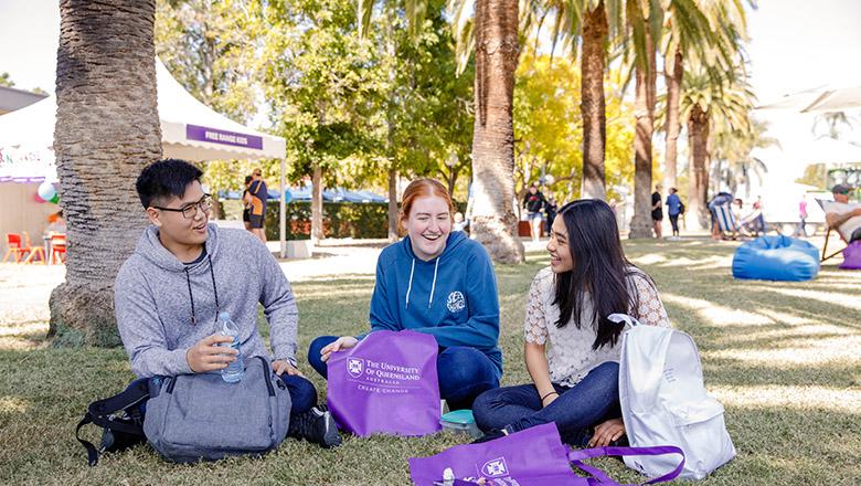 Three students sit on the grass under the palm trees at UQ's Gatton campus with their purple open day totes bags, chatting and laughing