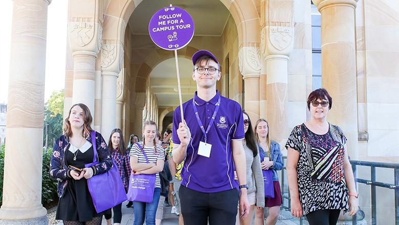 A student ambassador wearing a purple polo shirt holds a sign saying 'follow me for a campus tour' and leads a group of people through UQ's sandstone cloisters