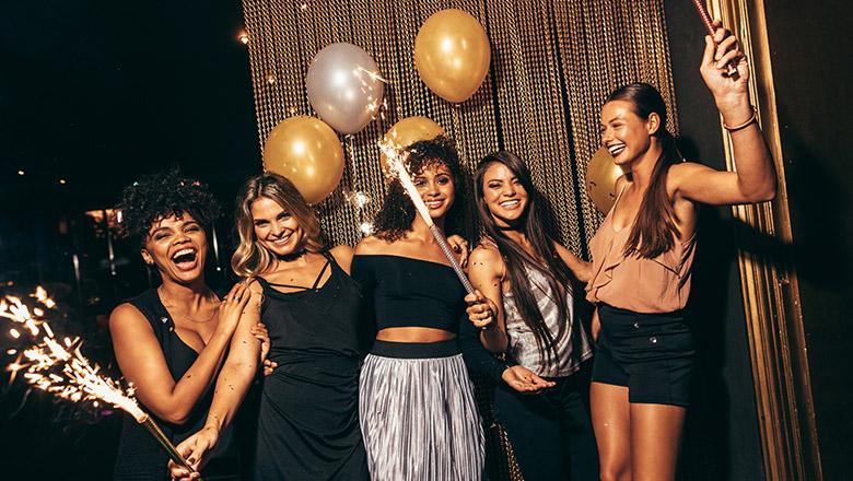 Group of girls dressed in party outfits post for a photo laughing and holding sparklers in front of balloons