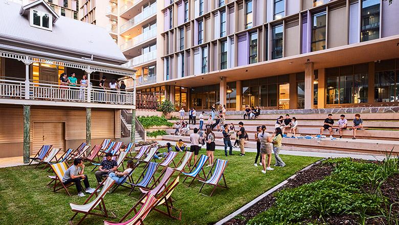 Stripy deck chairs are lined up on green grass with students mingling around them, a Queenslander in the background and a high rise building