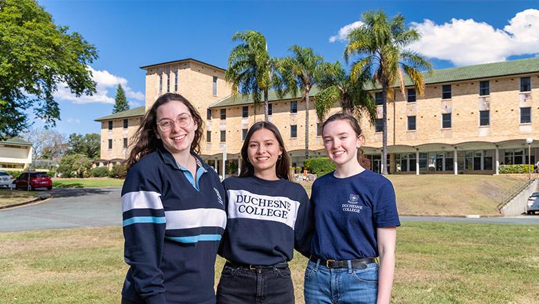 Three young women stand smiling in front of UQ's Duchesne College, with palm trees and building in background