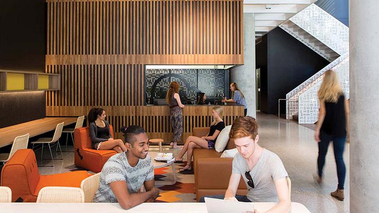 Students sit in the lobby of Iglu Brisbane City student accommodation on orange armchairs with wooden slat architecturally designed reception desk in background