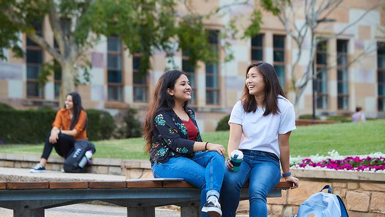 Two students sit on a bench chatting in front of UQ's iconic sandstone buildings