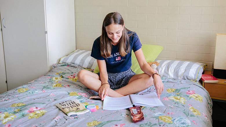 A young woman sits on her bed in a college dorm, reading a study printout