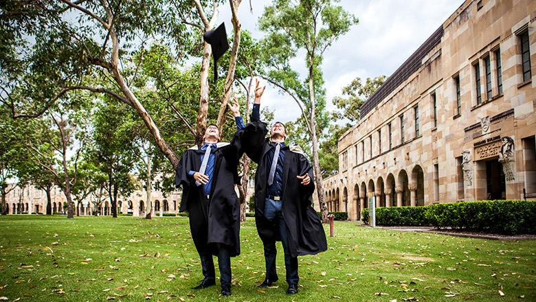Two UQ graduates stand in the Great Court in their graduation gowns, throwing their mortarboards into the air