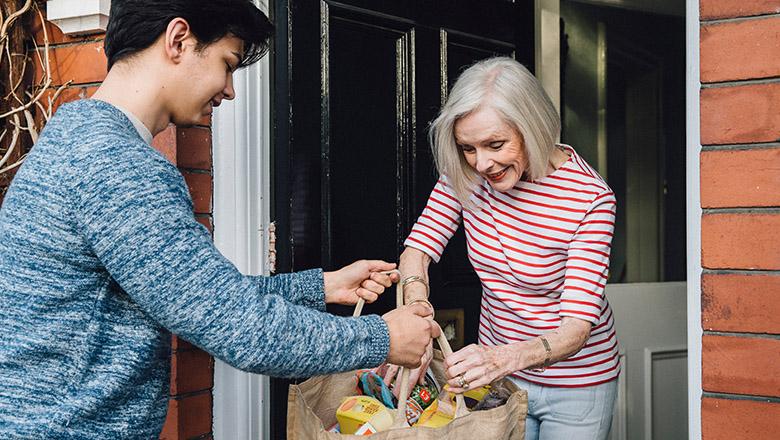 A teenage boy hands groceries to an elderly woman at her front door.