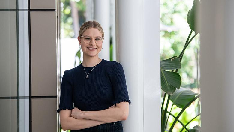 Rose Foster stands outside of UQ's Student Central leaning against a white pillar and smiling with plants in the background