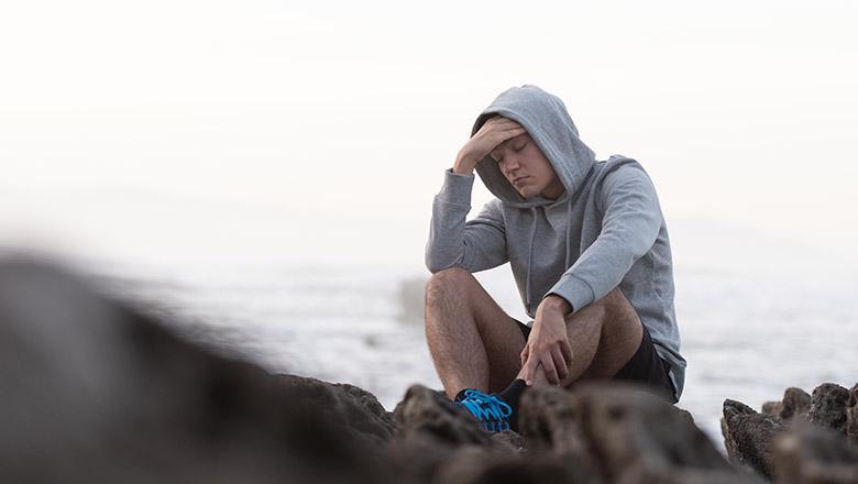A young man sits isolated on rocks in running gear, with the ocean in the background. His head is in his hand and his eyes are closed.
